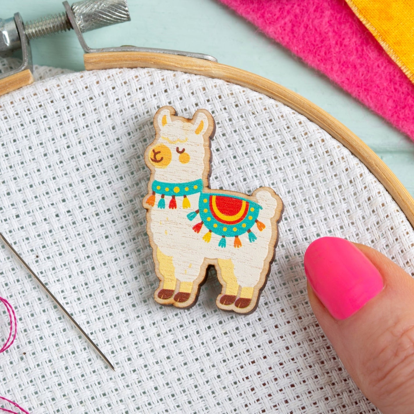 Embroidery hoop with a llama needle minder design, colourful threads, and a hand holding a needle on a light blue background.