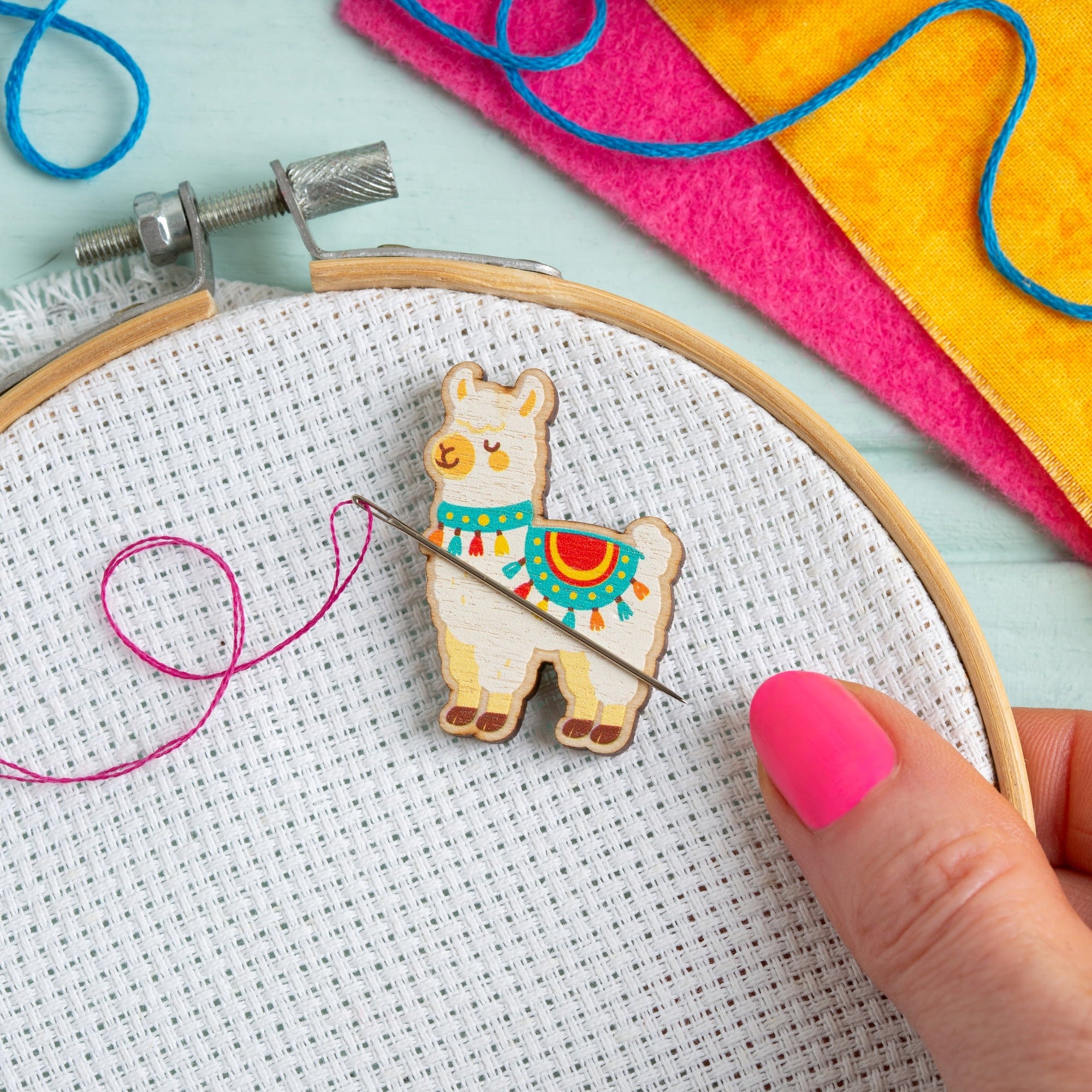 Embroidery hoop with a colorful Alpaca needle minder design, pink yarn, and a hand holding a needle on a light blue background.