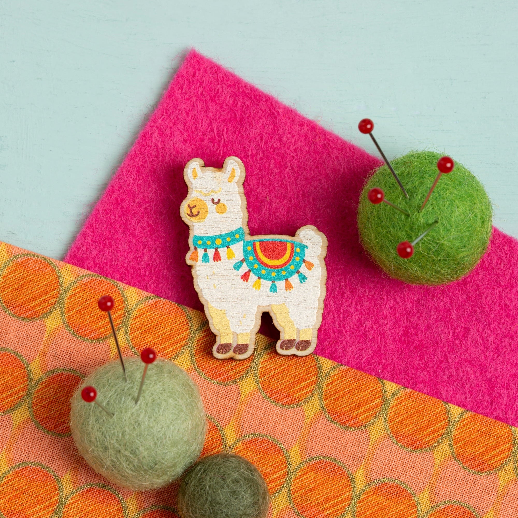 Alpaca-shaped needle minder on a colourful fabric background with felt balls and pins.