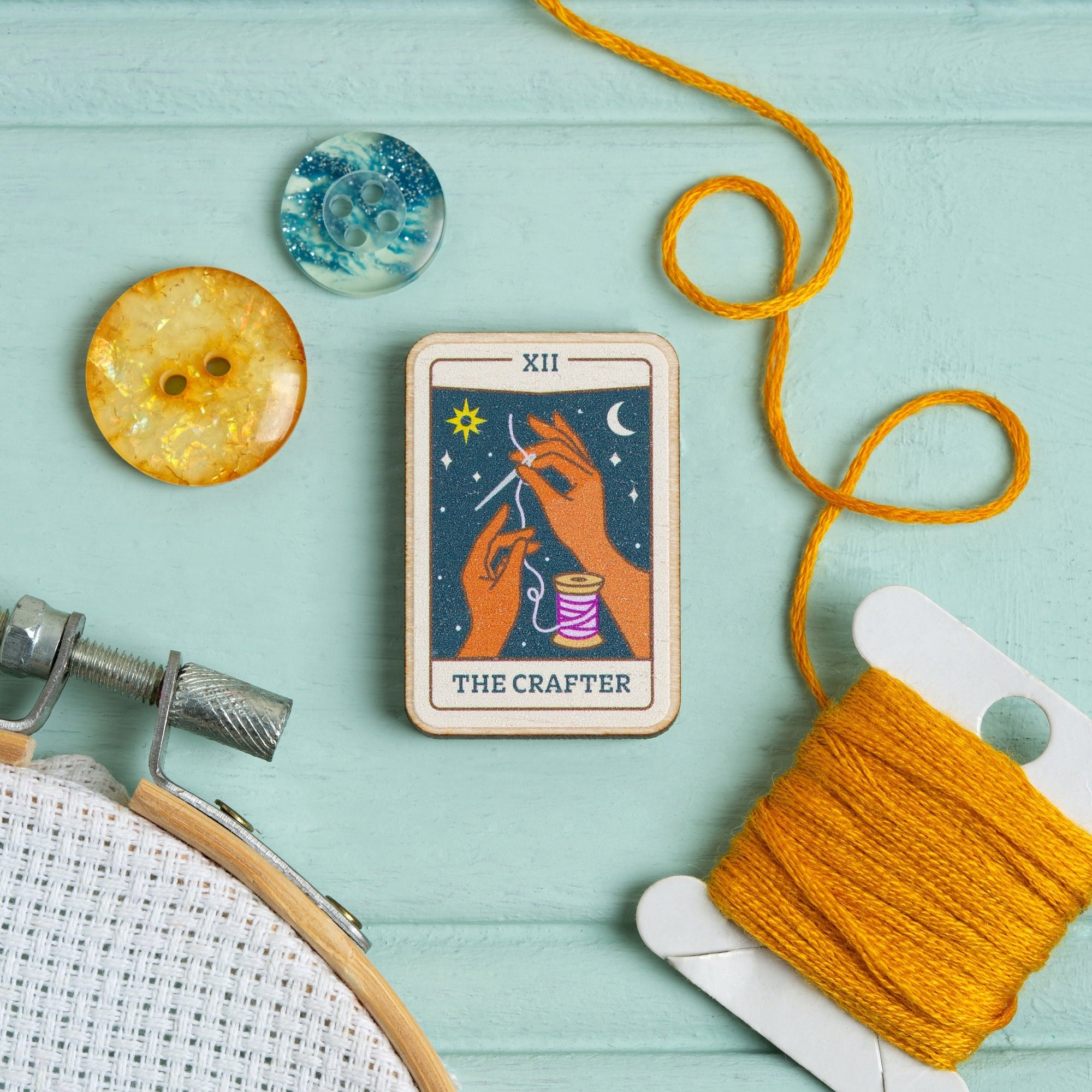 Tarot card needle minder titled 'The Crafter' with embroidery hoop, yarn, and dried orange on a teal background
