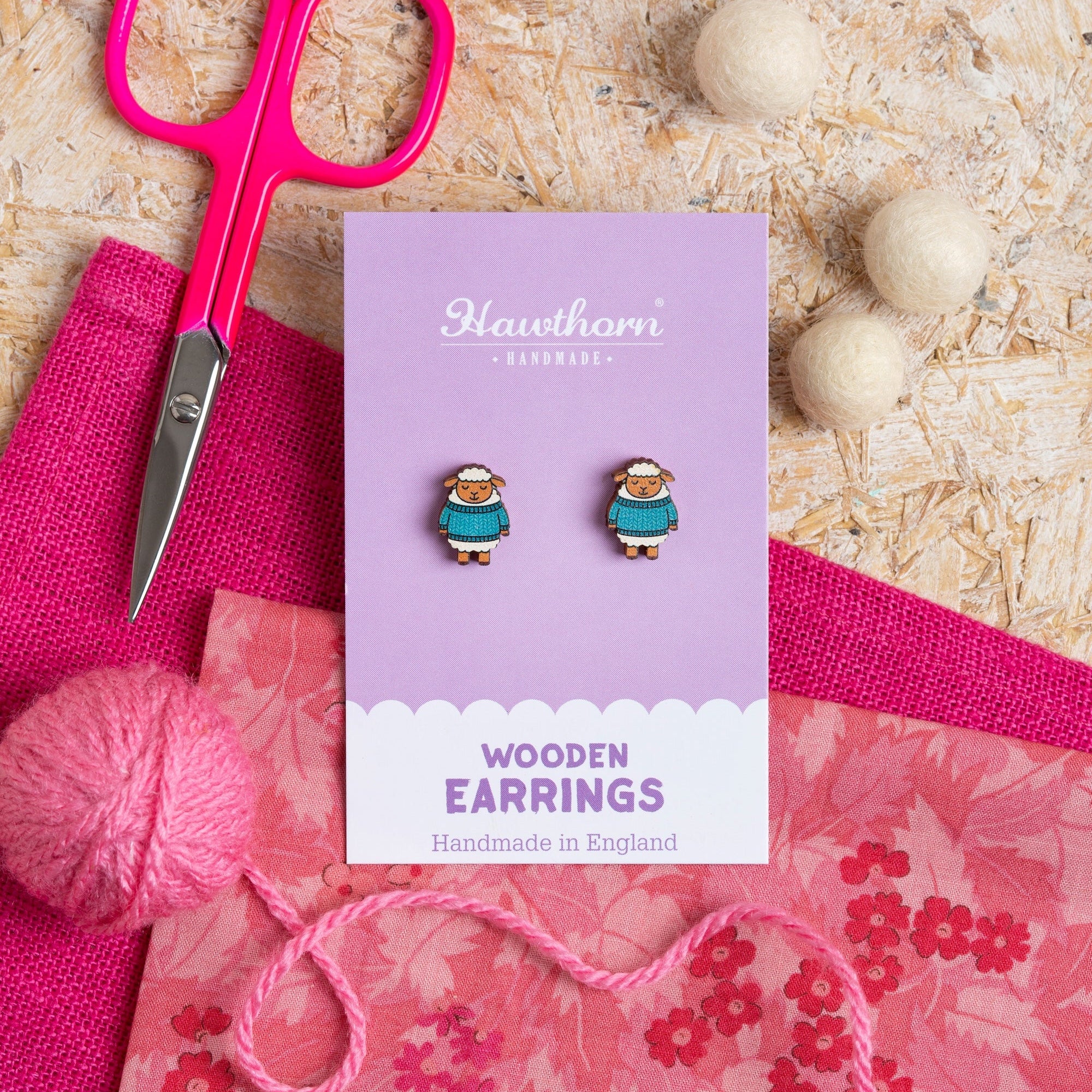 Woolly Sheep wooden earrings on a purple card with pink scissors and yarn on a wooden surface