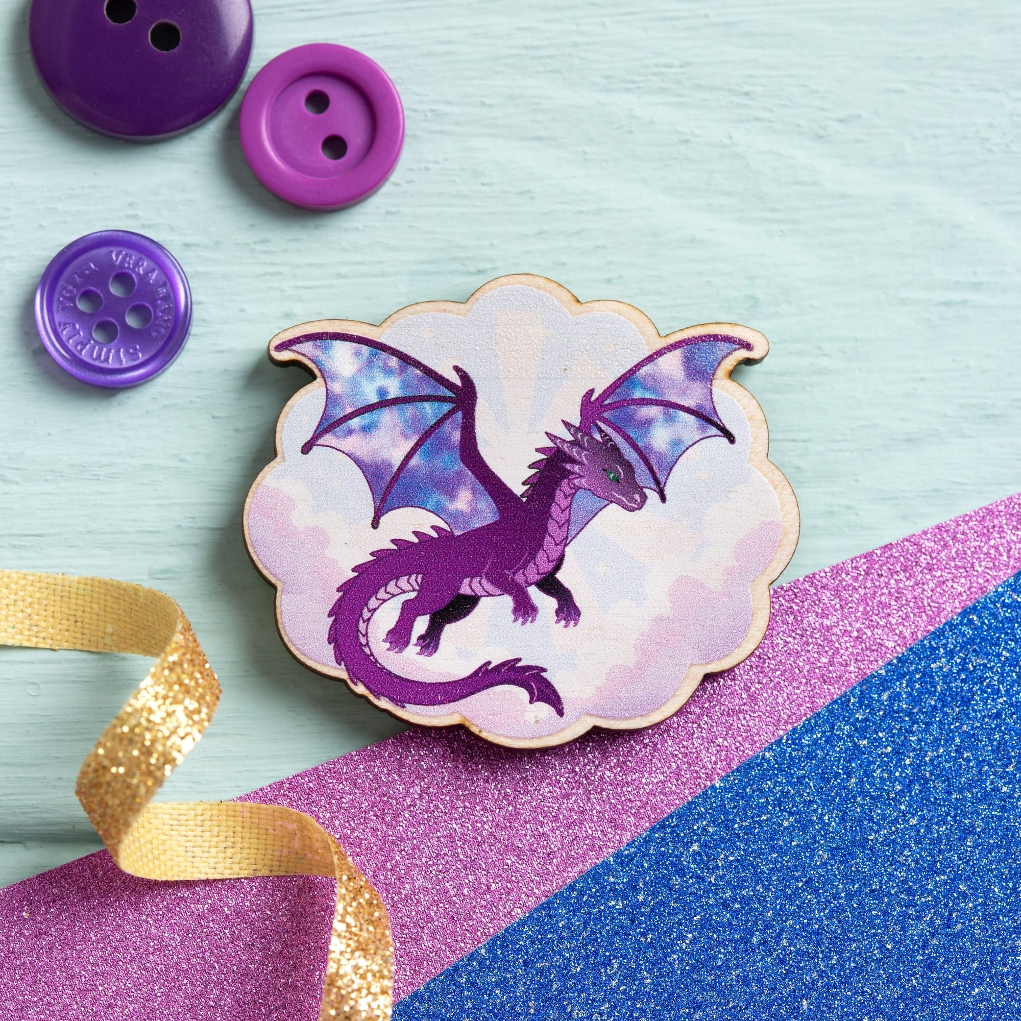 A dragon-themed cloud-shaped needle minder or badge in purple and pastel tones, styled on a light blue wooden surface with purple buttons, a gold ribbon, and pink and blue glitter paper.