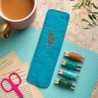 Teal embroidered owl bookmark with wooden spools of thread on a textured surface with a cup of coffee and pink scissors.