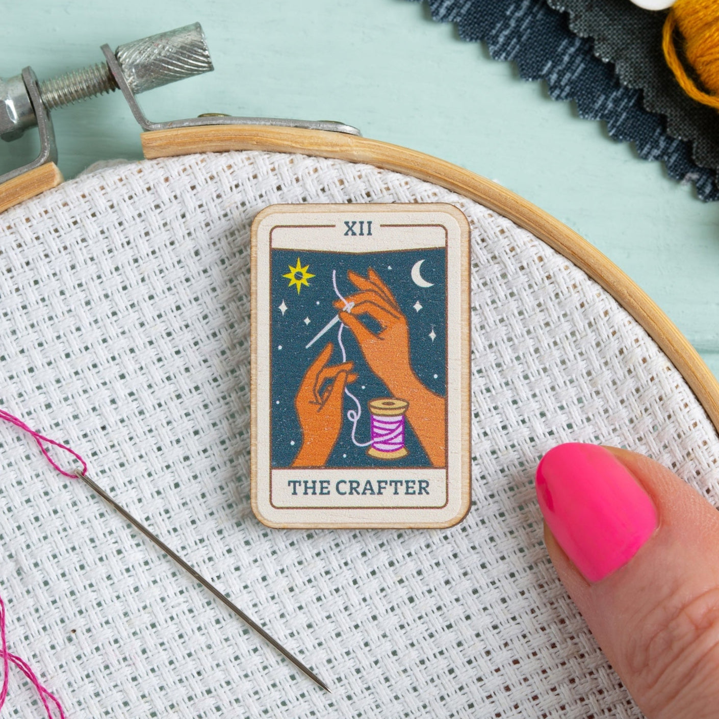 Embroidery hoop with a tarot card needle minder labeled 'The Crafter' on fabric.