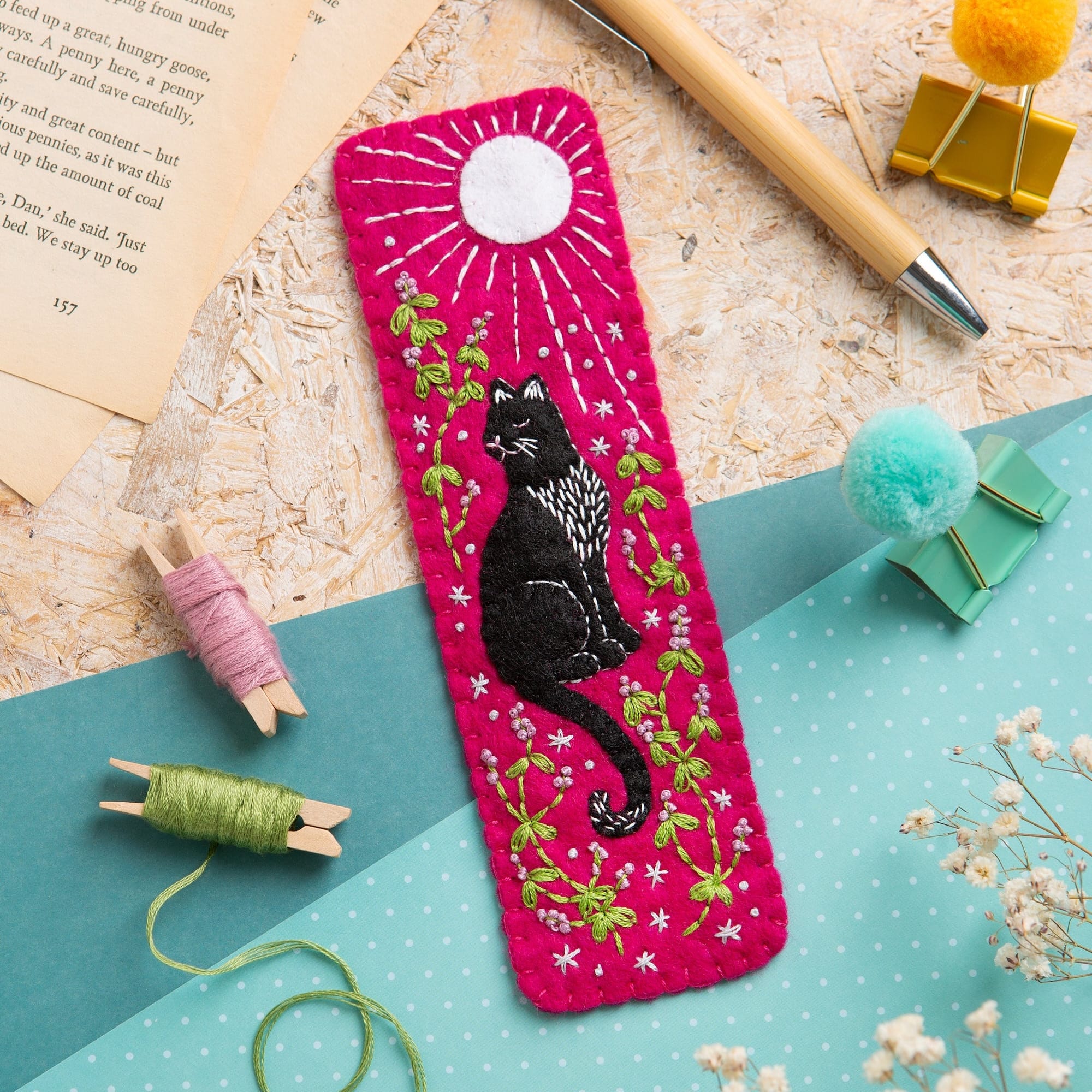 A bright pink rectangular fabric bookmark featuring an embroidered black cat, green leaves, and white flowers under a white sun motif. The bookmark is styled in a flat-lay arrangement on a light blue polka-dot surface alongside a wooden pen, book pages, yarn, and small pom-poms.