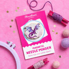 A Needle Minder dragon is displayed in a styled flat-lay on a pink background, surrounded by craft supplies including a white embroidery hoop, purple and pink thread wraps, and pom-poms.
