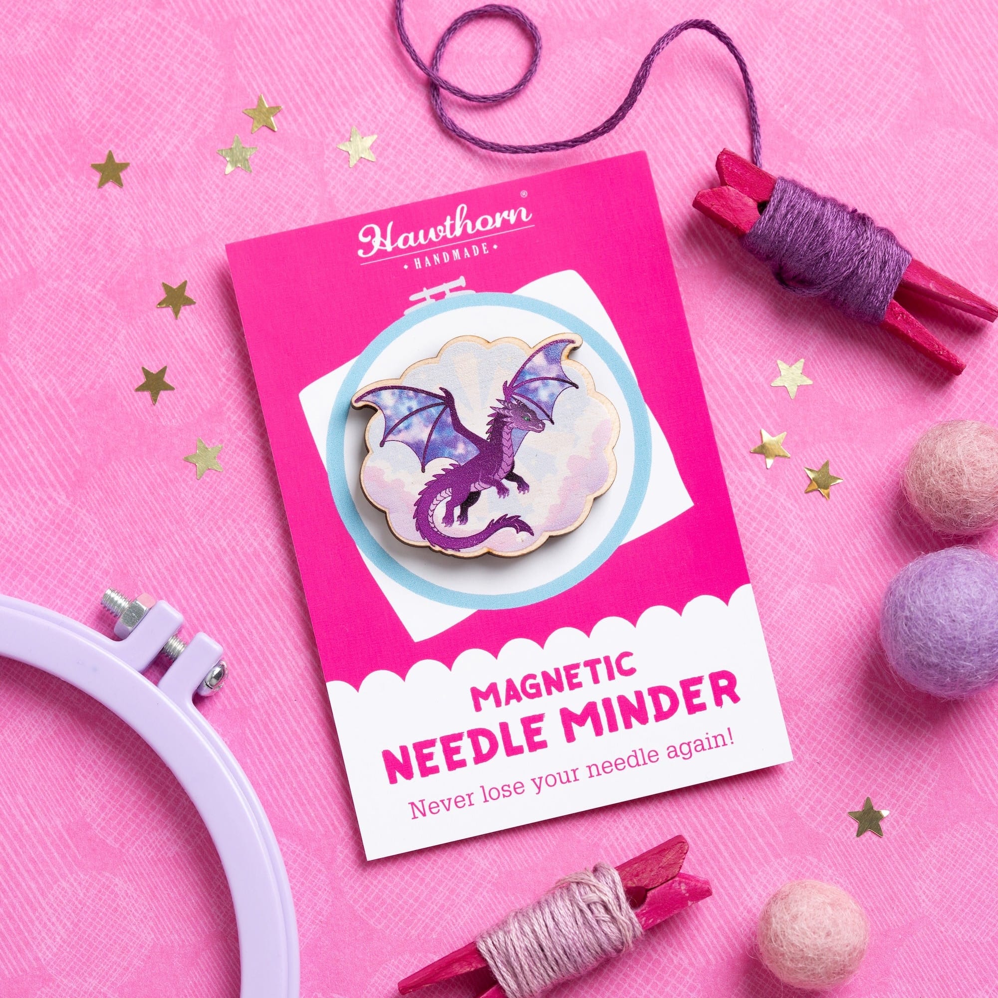 A Needle Minder dragon is displayed in a styled flat-lay on a pink background, surrounded by craft supplies including a white embroidery hoop, purple and pink thread wraps, and pom-poms.