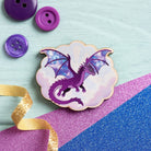 A dragon-themed cloud-shaped needle minder or badge in purple and pastel tones, styled on a light blue wooden surface with purple buttons, a gold ribbon, and pink and blue glitter paper.