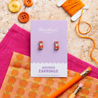Fox Wooden earrings on a card with orange thread, buttons, and a pink cloth on a textured surface.