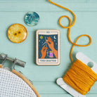 Tarot card needle minder titled 'The Crafter' with embroidery hoop, yarn, and dried orange on a teal background