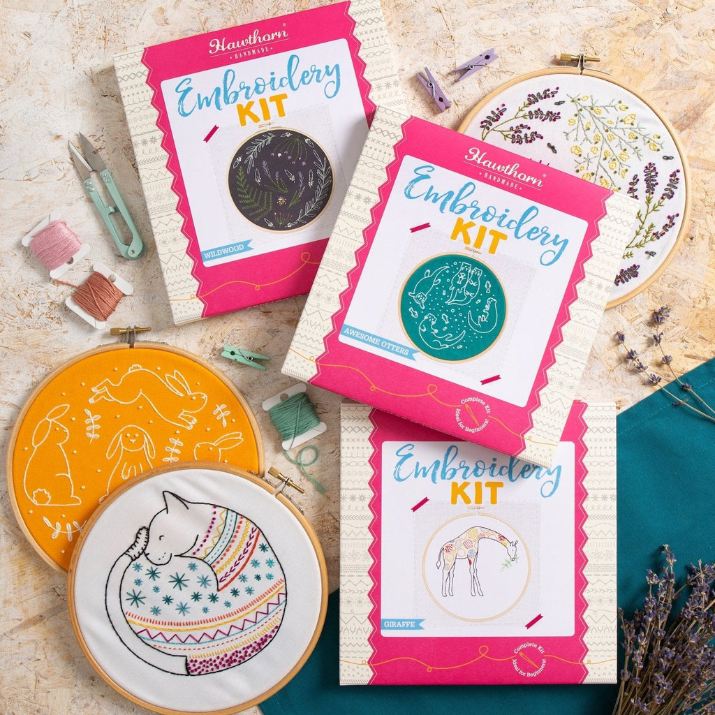 Group image of three completed embroidery kits and three embroidery kit boxes displayed on wooden background.