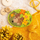 second image of hedgehog felt craft brooch displayed on wooden and mustard colourred background with yellow twine and yellow painted pine cones.