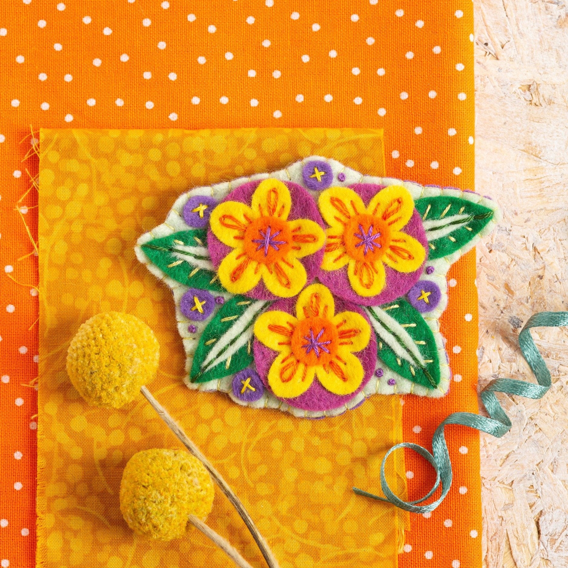 Penelope flower brooch displayed on orange fabrics on a wooden background.