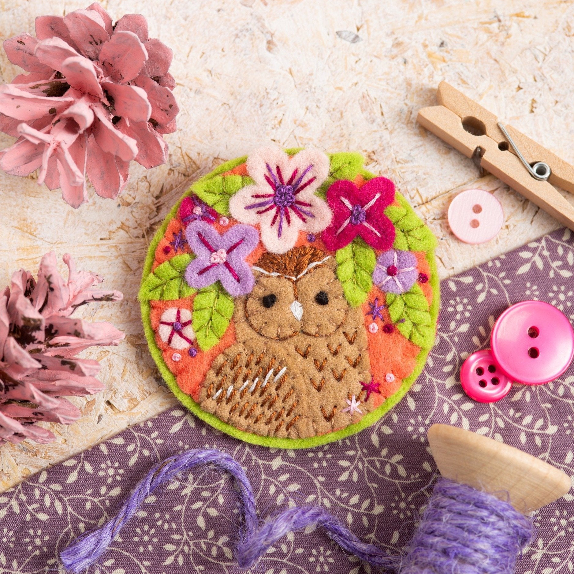 Tawny owl brooch on wooden background with purple fabric, wooden pegs and pink painted pine cones used as props.