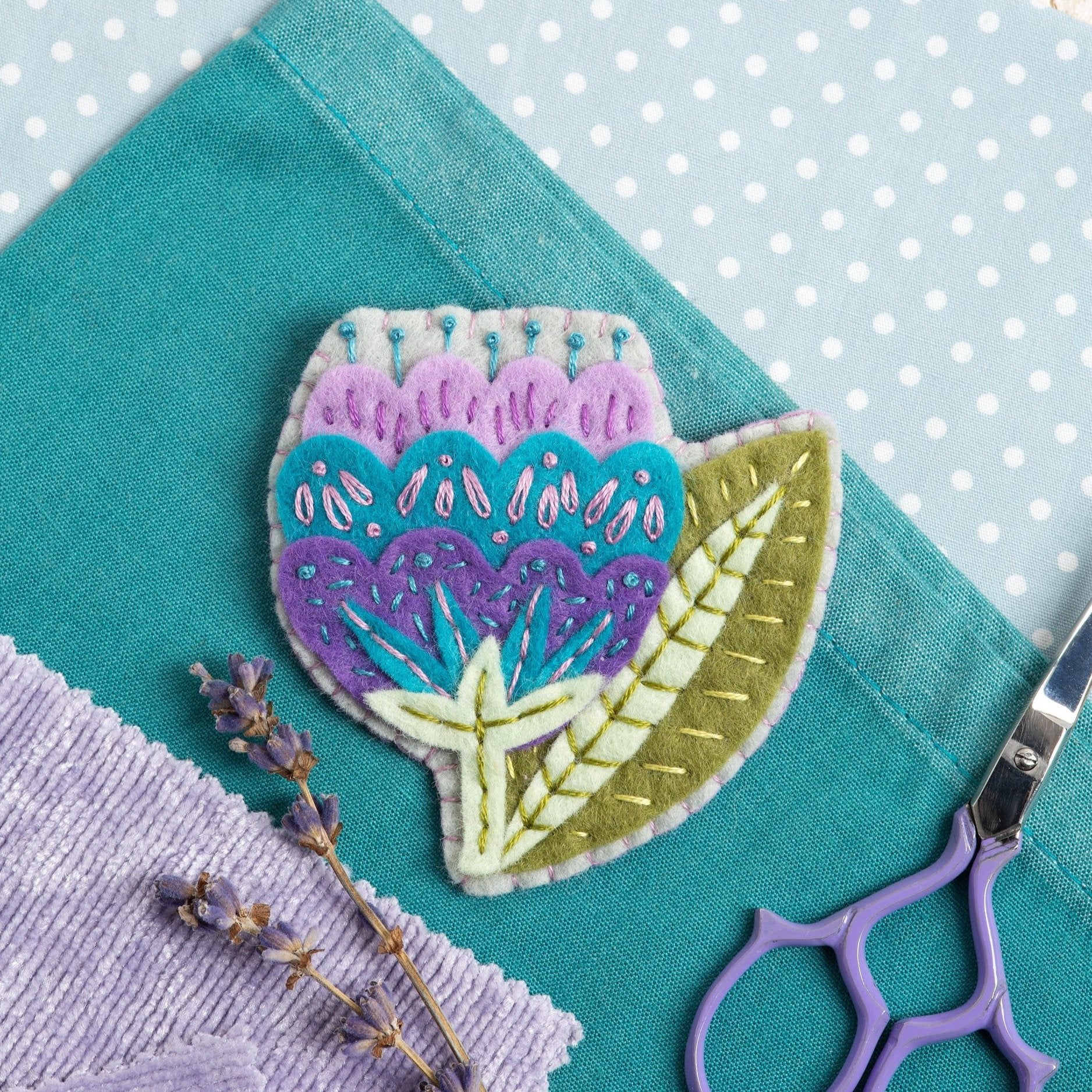 Vita flower felt craft brooch kit displayed on pale blue polka dot background, teal and lilac fabric squares and purple scissors.
