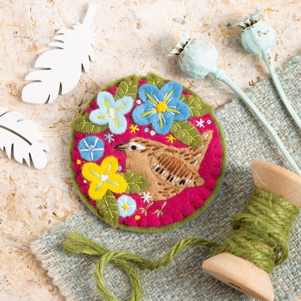Wren felt craft brooch on wooden background with green thread on wooden spool displayed next to it.