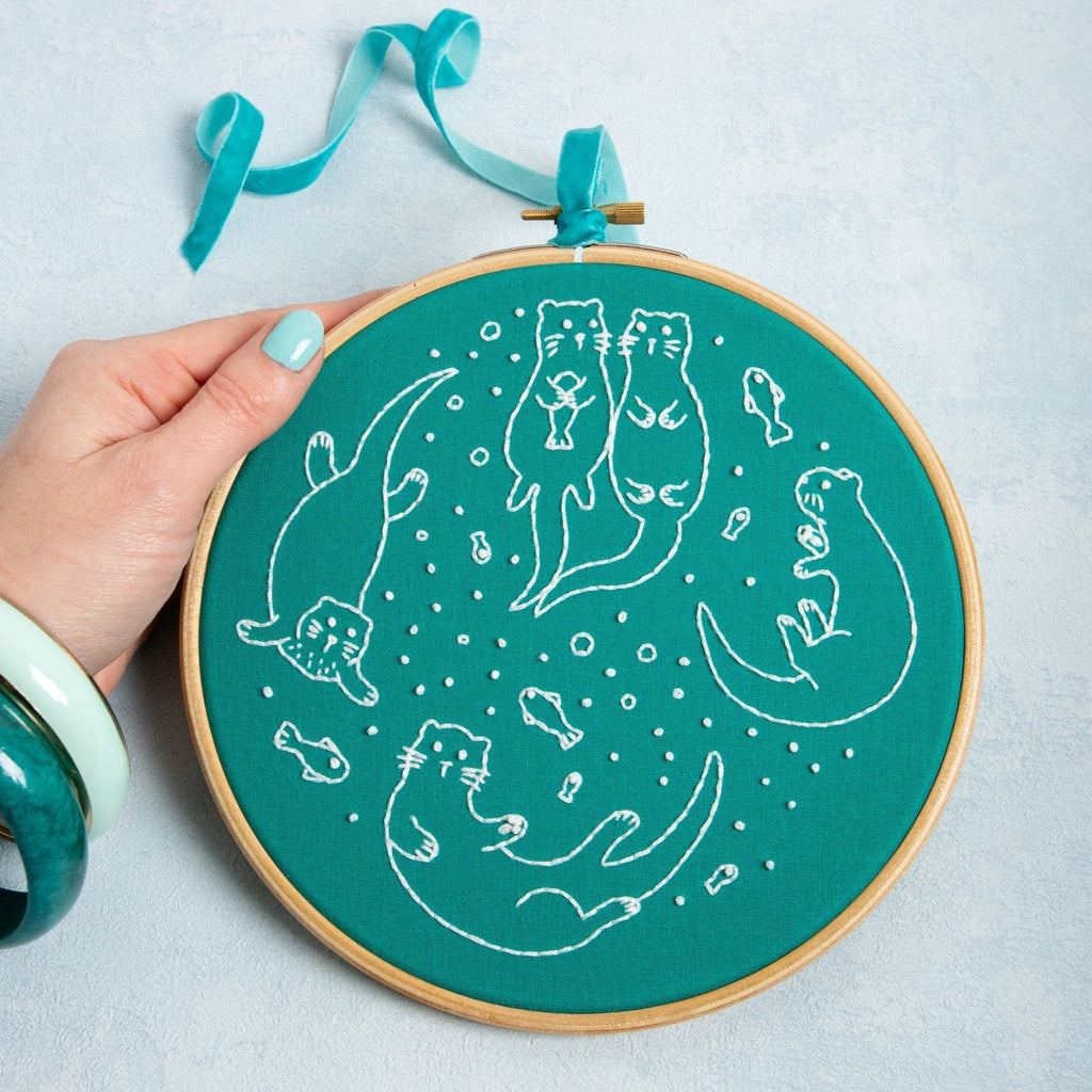 Completed Awesome Otters Embroidery Kit being held up by hand with mint green painted fingernail.